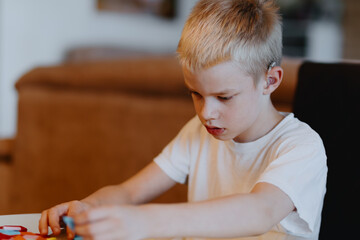 A boy with hearing aids focuses intently while playing with building blocks and board games,...