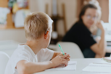 A boy with hearing aids focuses on homework, showcasing natural inclusive education. Authentic...