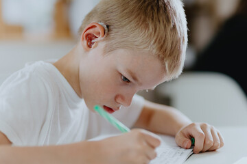 A boy with hearing aids focuses on homework, showcasing natural inclusive education. Authentic...