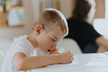 A boy with hearing aids focuses on homework, showcasing natural inclusive education. Authentic...