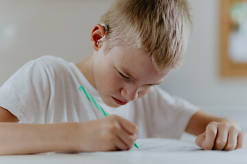 A boy with hearing aids focuses on homework, showcasing natural inclusive education. Authentic...