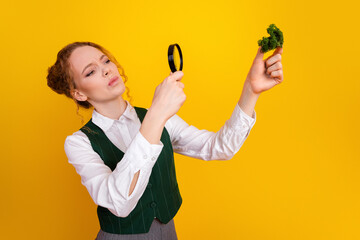Focused young woman examines fresh kale closely using a magnifying glass against a yellow background