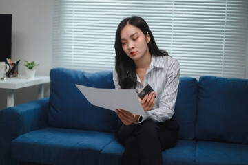 Asian businesswoman working from home is sitting on sofa comparing information on smartphone and...
