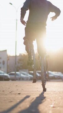 Back shot of guy wearing gray hoodie and dark gray pants jumping on spring stilts on paved road in residential area surrounded by houses during sunset, showcasing dynamic motion