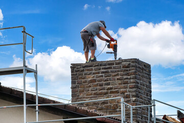 Worker with jackhammer starts demolishing a brick chimney on the roof of a residential building