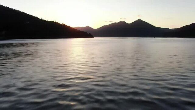 Aerial flight over calm lake towards silhouetted mountains at sunset.
