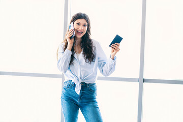 Cheerful young woman holding a passport and talking on the phone at a bright airport terminal,...