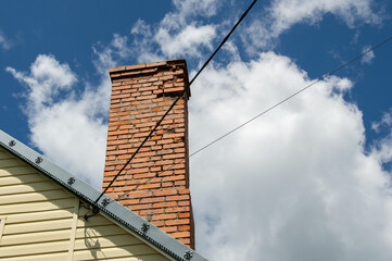 A tall brick chimney stands on a house roof under a bright blue sky with scattered white clouds,...