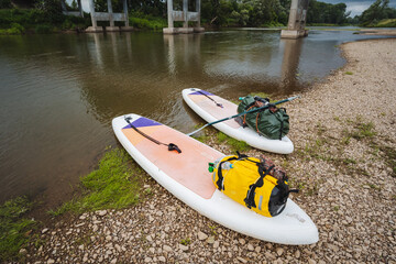 Two vibrant paddle boards are resting peacefully on the sandy shore of a beautiful river, waiting...