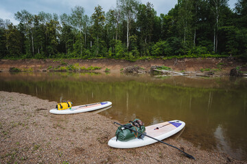 Two paddle boards are currently resting on the sandy shore of a picturesque river, waiting for...