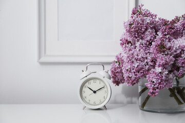 Beautiful lilac flowers and alarm clock on table near white wall indoors, closeup