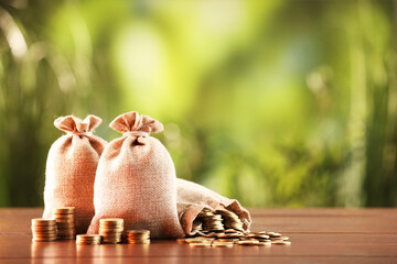 Burlap bags and stacked coins on wooden table against blurred background, space for text