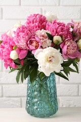 Bouquet of beautiful peonies in vase on white table against brick wall indoors, closeup