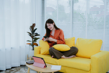 Young Asian woman holding smartphone playing social media, sitting on sofa in living room at home....