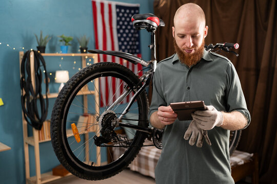 Caucasian bearded man repairing bicycle at home using digital tablet repairing and servicing bikes in workshop - Powered by Adobe