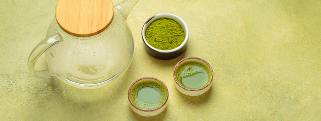 Steaming glass teapot with wooden lid beside two small cups of matcha tea and a bowl of matcha...