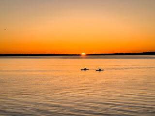 Silhouettes of kayakers paddling across peaceful sea as the sun sets behind the distant horizon