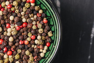 Mixed peppercorns in green bowl. Colorful dried red black white and green pepper seeds on dark stone background. Aromatic spice mix closeup. Culinary seasoning texture for cooking or restaurant menu.