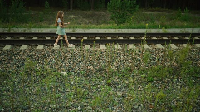 Side view of little girl walking beside railway sleeper holding grass strand in hand, wearing denim dress and striped shirt, surrounded by gravel stones and wild plants