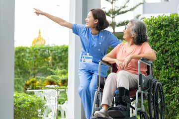 smiling Asian caregiver in scrubs enjoys a moment outdoors with senior woman in a wheelchair,...