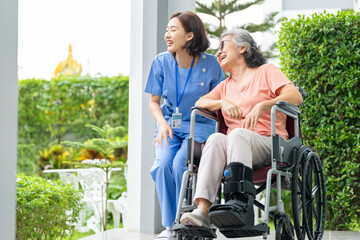 smiling Asian caregiver in scrubs enjoys a moment outdoors with senior woman in a wheelchair,...