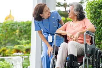 smiling Asian caregiver in scrubs enjoys a moment outdoors with senior woman in a wheelchair,...