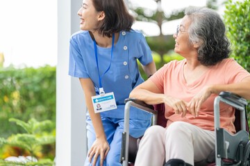 smiling Asian caregiver in scrubs enjoys a moment outdoors with senior woman in a wheelchair,...