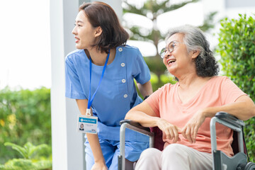 smiling Asian caregiver in scrubs enjoys a moment outdoors with senior woman in a wheelchair,...
