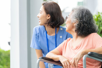 Obraz premium smiling Asian caregiver in scrubs enjoys a moment outdoors with senior woman in a wheelchair, pointing at nature enthusiasm. Concept of home healthcare, assisted living, and positive elderly support.