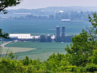 Farmland with silos, barns, and houses shimmers in a summertime haze, seen from an outlook above,...