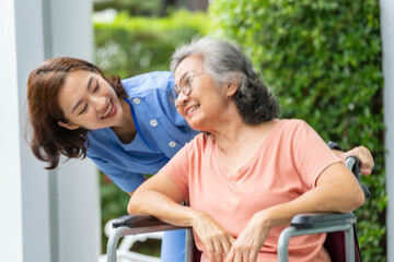 smiling Asian caregiver in scrubs enjoys a moment outdoors with senior woman in a wheelchair,...