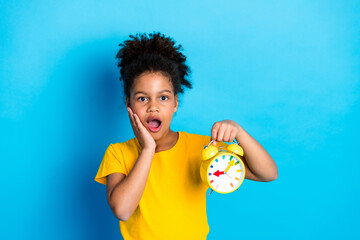 Surprised young girl with curly hair in yellow t-shirt holding colorful alarm clock in hand against blue background