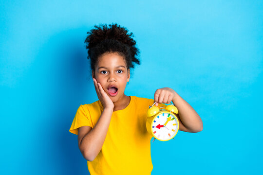 Surprised young girl with curly hair in yellow t-shirt holding colorful alarm clock in hand against blue background - Powered by Adobe