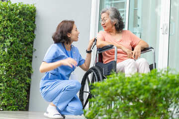 smiling Asian caregiver in scrubs enjoys a moment outdoors with senior woman in a wheelchair,...