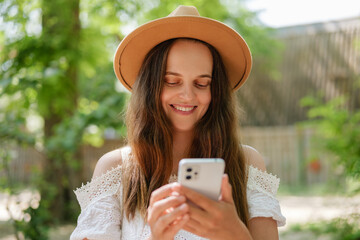 A smiling young woman in a tan hat and white lace top looks at her phone outdoors. Surrounded by...