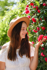 A smiling young woman in a brown hat and white dress gently touches blooming red roses in a vibrant...