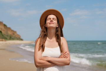 
A serene woman in a white dress and hat stands on a sunny beach with her eyes closed and arms...