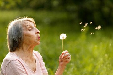 A close-up portrait of a senior woman blowing on a dandelion, sending its seeds floating into the...