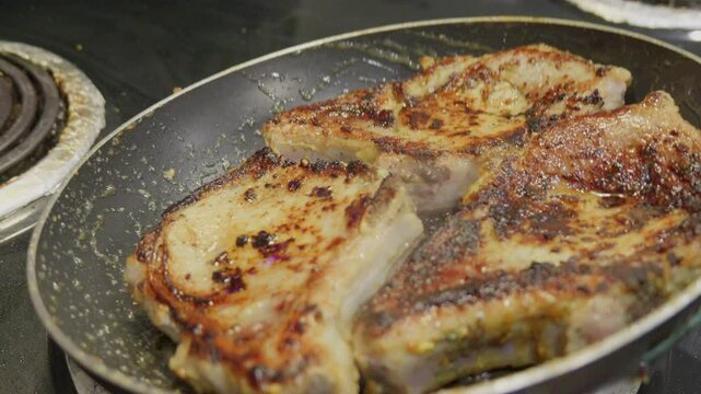 Close-up of a hand using chopsticks to flip sizzling pork chops in a frying pan. The meat is browned and juicy, capturing the essence of home cooking