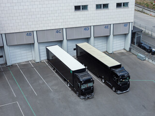 Two large black semi-trucks are parked at a loading dock with numbered bays in an industrial area,...