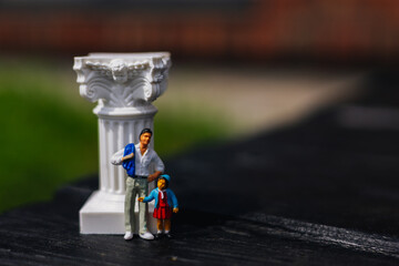 A miniature father and daughter stand beside a decorative pillar outdoors. The figures of a dad and...