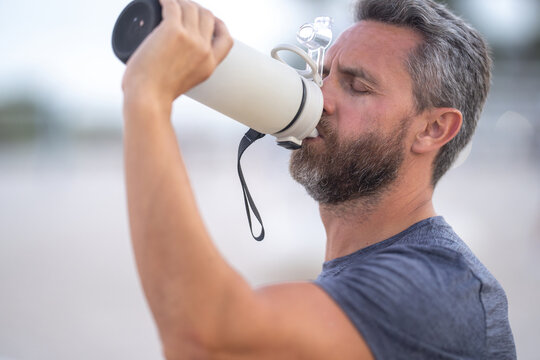 Sport fitness man resting on a bench and drink water from sport water bottle. Mature sport model sitting on a bench after a workout. Man taking break after fitness drink water from sport water bottle.