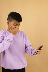 Vertical image of a young brown skinned Latino man wearing a lilac sweatshirt with a horrified expression with his hand over his mouth when seeing something on his cell phone on a brown background