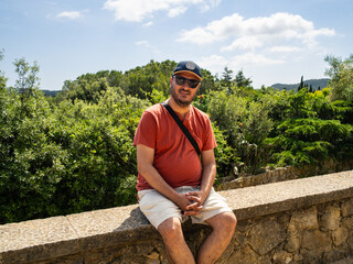 Man poses on a stone wall during holidays surrounded by trees under sunny skies looking at camera....