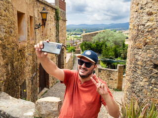 A man enjoys a sunny day in a charming stone village, posing for a selfie while giving a peace...