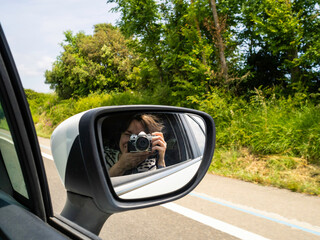 A person sitting in a car takes a self portrait through the side mirror. Lush greenery lines the...