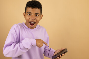 Cheerful young Latino man wearing a lilac sweatshirt pointing at his cell phone on a light brown background with space to write advertising, promotion and offer concept