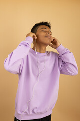 Vertical image of a latin american teenager wearing a lilac sweatshirt with his eyes closed, feeling the rhythm, listening to music with wired headphones on a light brown background with copyspace