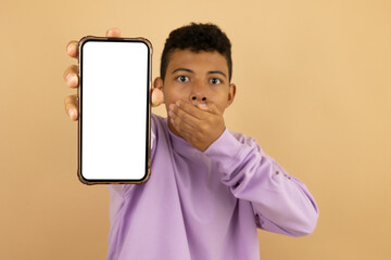 Latino teenager wearing a purple sweatshirt with a surprised expression covering his mouth, holding his cell phone with his hand with a blank screen with advertising space on a light brown background