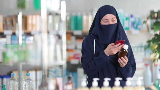 Focused young female customer of European appearance in traditional Muslim abaya and khimar(niqab) in pharmacy scanning qr code on bottle of medicinal syrup using phone to check usage or composition